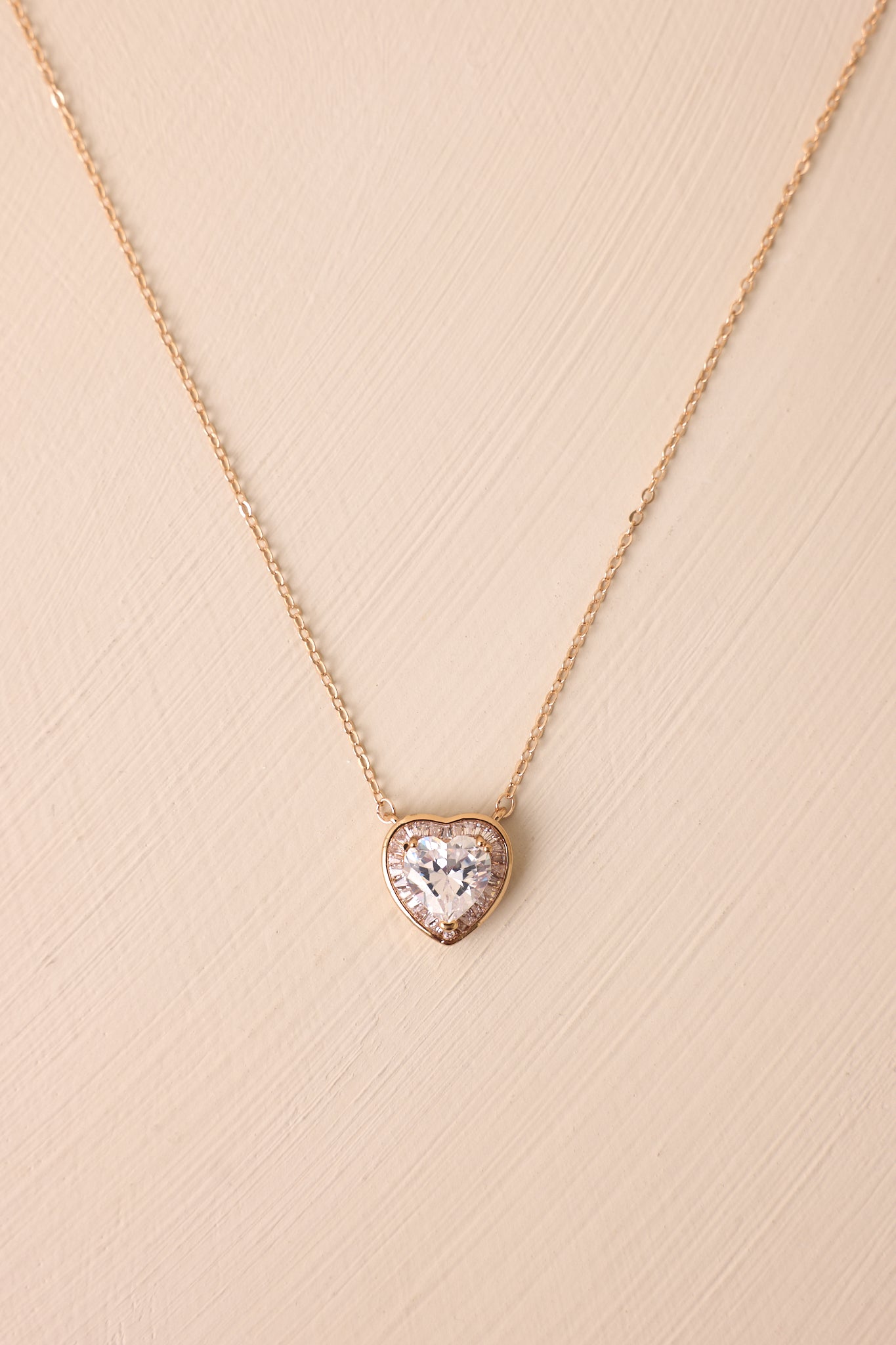 A closer look at the gold necklace featuring a thin gold chain and a heart-shaped rhinestone pendant, surrounded by a second layer of rhinestones and a gold border for added sparkle