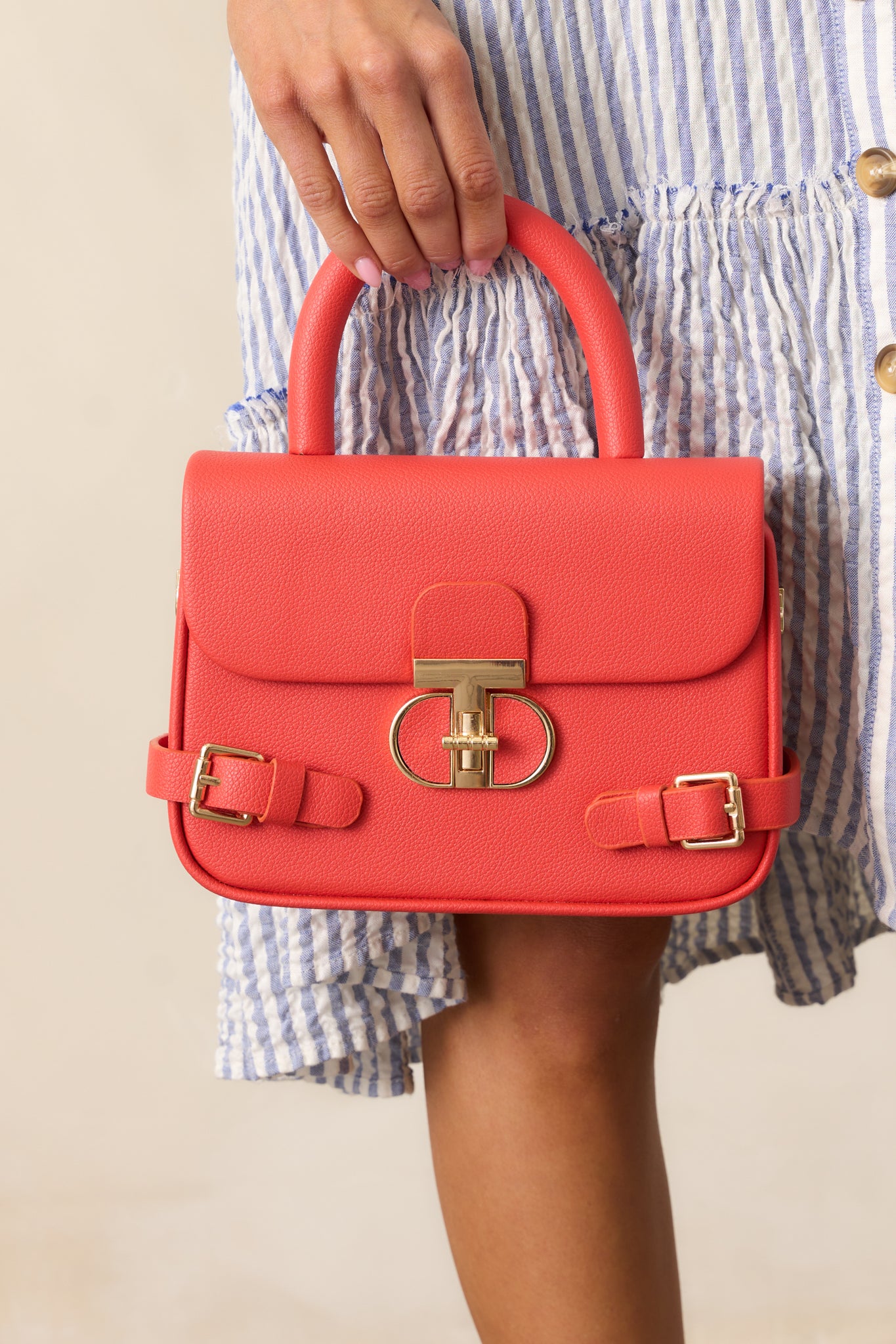 A full view of the burnt orange handbag featuring a rounded square shape, gold hardware, fold-over closure with a gold twist lock, and a red handle.