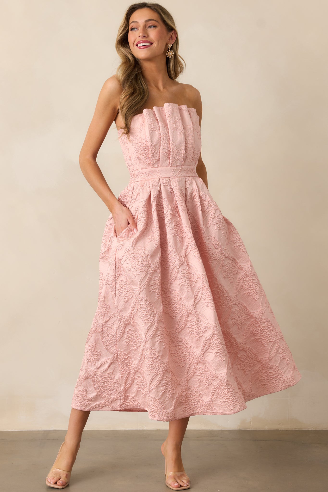 A angled side view of the blush dress emphasizing its strapless design, pleated bodice, and the smooth pleats along the skirt.