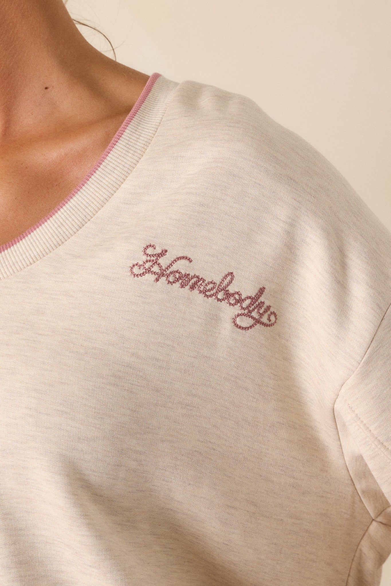 A detailed close-up of the "Homebody" embroidery on the upper left bust, with focus on the v-neckline, ribbed bottom hemline, and pink trim detailing on the sweatshirt.