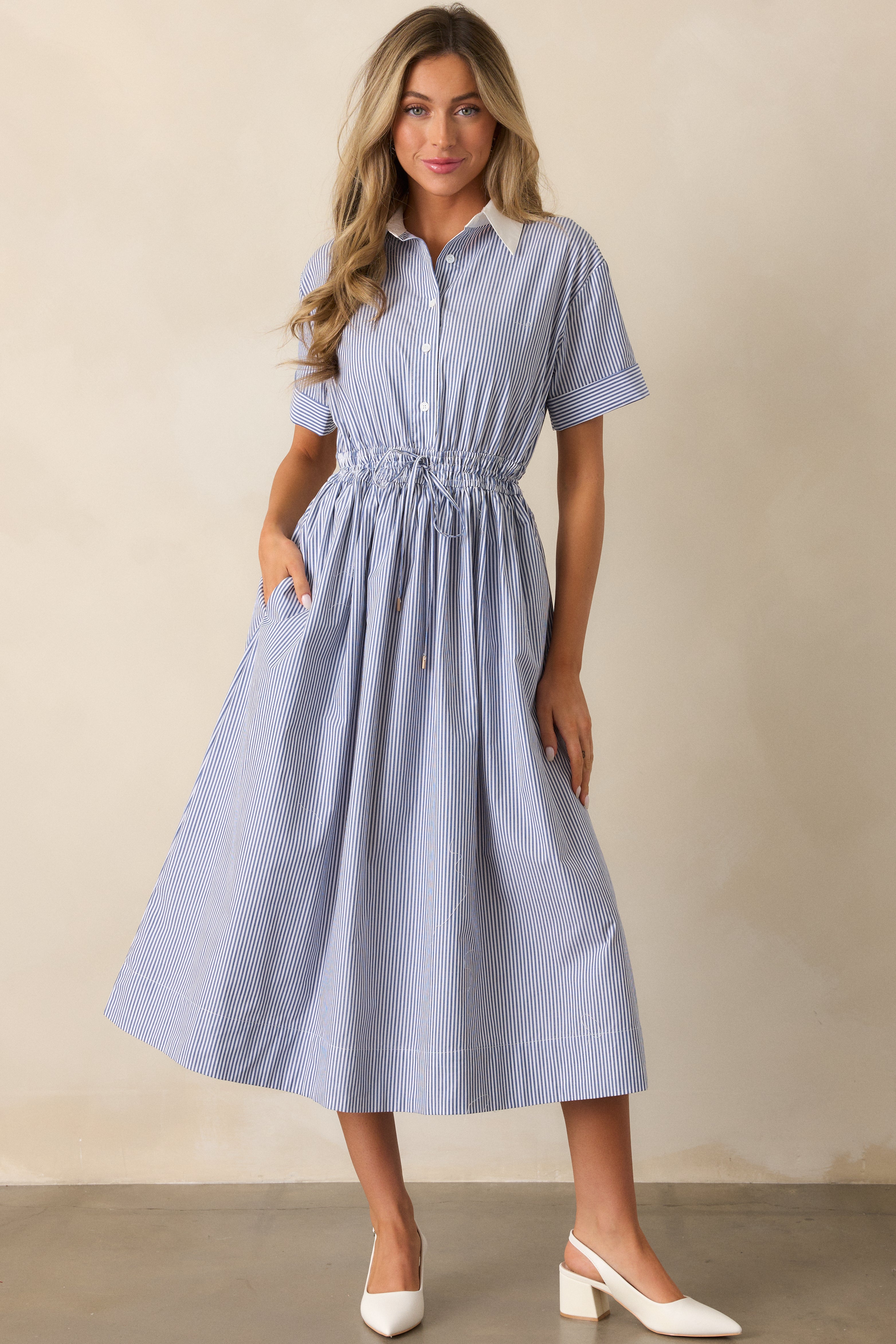 Full view of a blue dress featuring two functional pockets, a white collared neckline, short cuffed sleeves, and a fitted bodice.