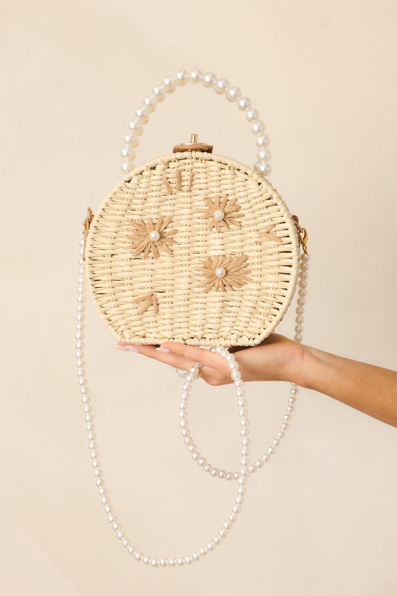 Ivory handbag in a circular shape, showcasing tan rattan flower accents with pearl centers, gold twist and lock closure, large faux ivory pearl beaded handles, and an optional faux pearl shoulder strap.