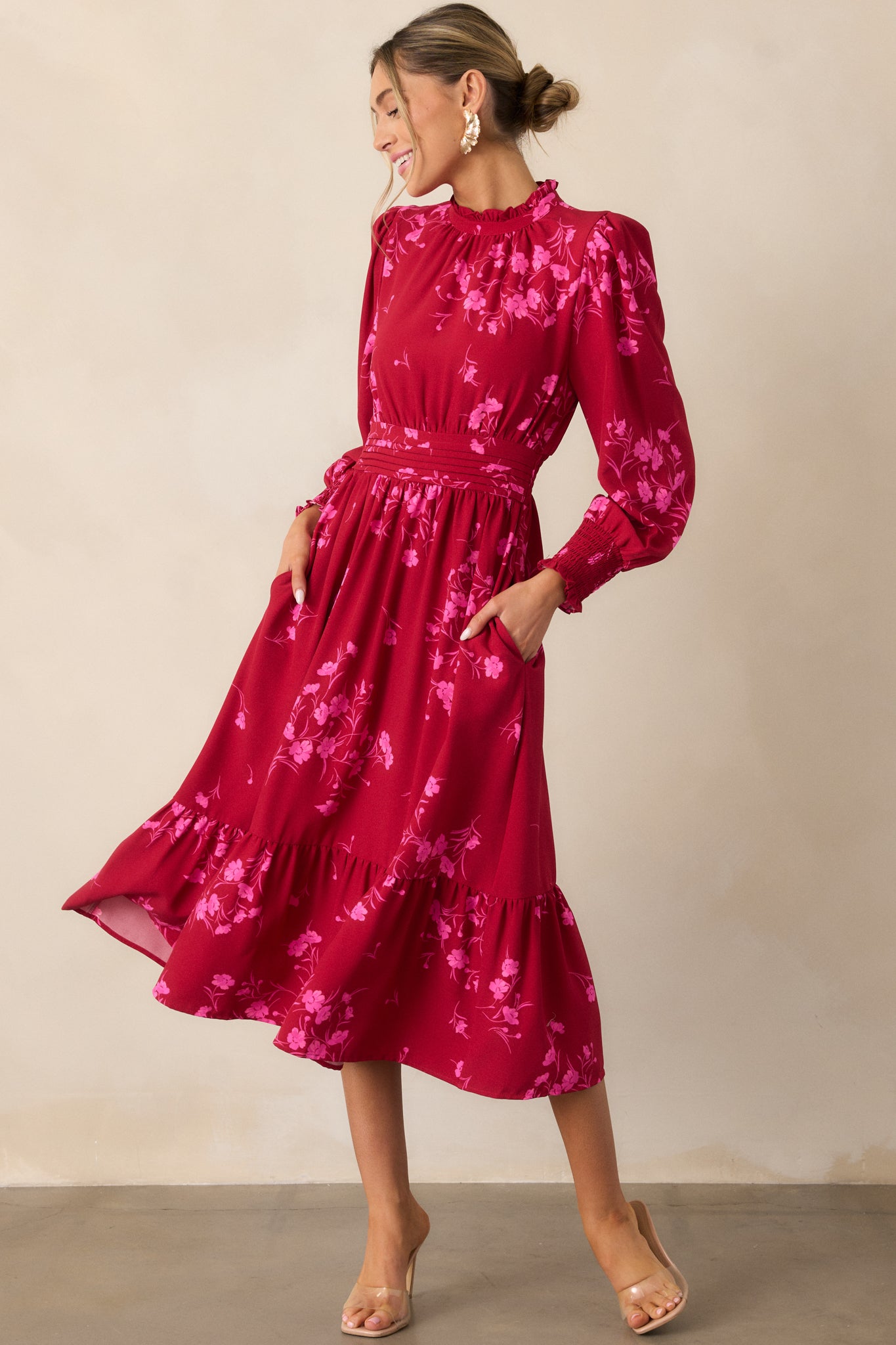  Showcasing the pink and red floral dress with its high ruffle detail neckline, pleated waistband, and long smocked sleeves, the dress cascades into a tiered, midi-length skirt that adds movement and charm.