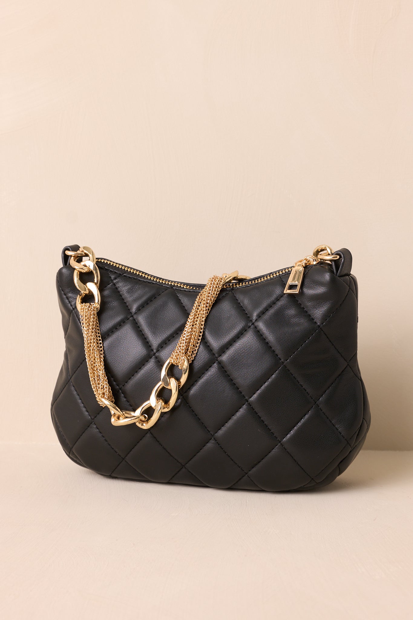 A side angle of the black faux leather handbag on a beige background, showing the quilted pattern and the structured silhouette with the thin gold chain shoulder strap resting nearby.